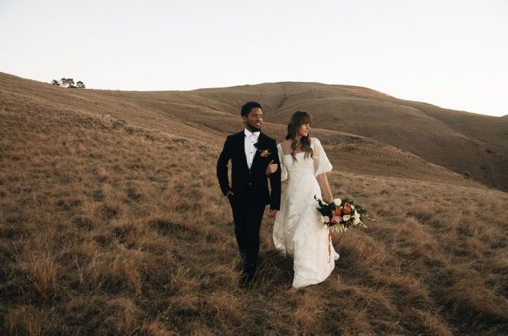 Amour Weddings Wild Hearts Wedding Vendor Directory - Connor and Sarah offer Photography and Film packages for the ultimate wedding experience, their style is about capturing raw and honest moments between people - bride and groom walking holding hands in a field